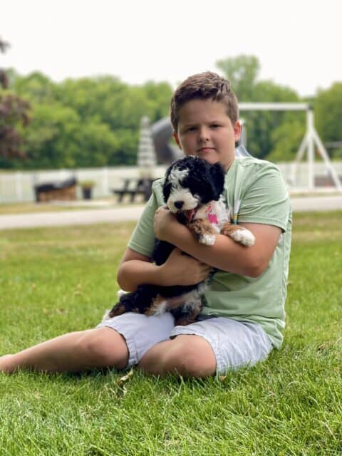 9-year-old boy holding a tiny Bernedoodle puppy in green yard