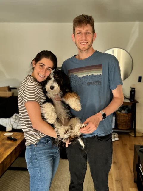 A couple holding their mini Bernedoodle between them while standing in their living room