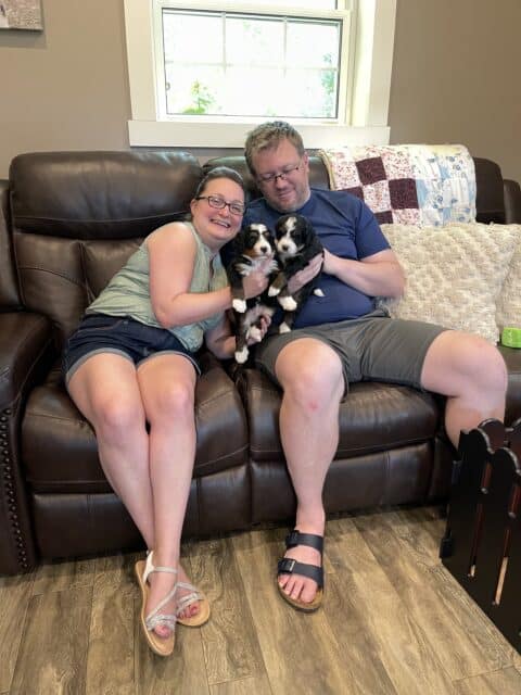 A husband and wife each holding a small Bernedoodle puppy while sitting on a couch at Fawn River Doodles