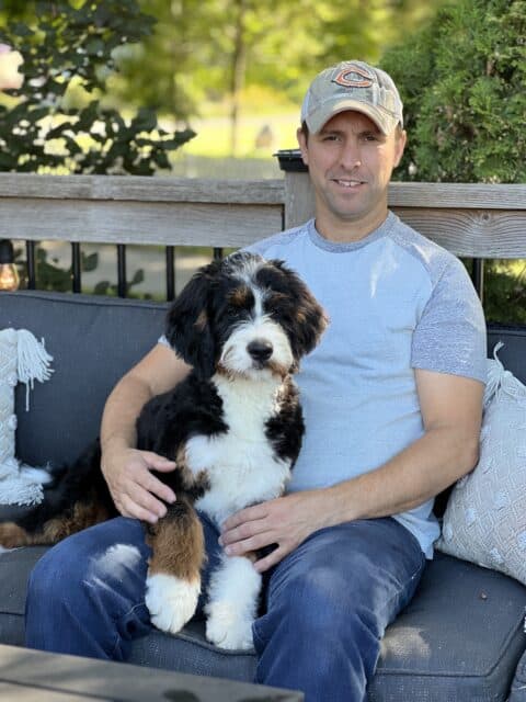 Robin holding medium-size, wavy-coat Bernedoodle in outdoor patio