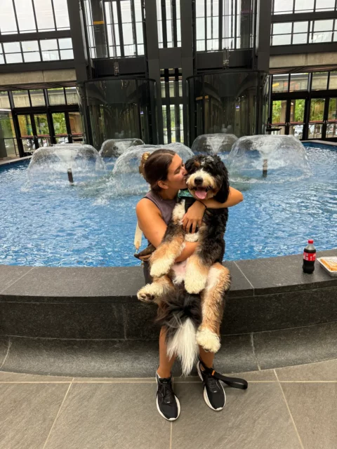 Woman kissing her Bernedoodle puppy while sitting on edge of a water fountain